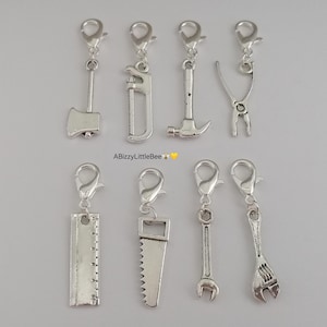 May include: Set of ten silver-toned metal charms shaped like various tools, including a hammer, saw, wrench, pliers, and ruler. The charms are attached to silver-toned lobster claw clasps.