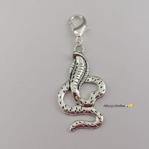May include: A silver cobra charm with a lobster clasp. The snake charm features scale detailing and a raised hood. The charm is designed for attachment to a bracelet or necklace. The text "ABizzyLittleBee" is visible in the image.
