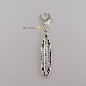 May include: Silver surfboard charm with a lobster clasp. The charm features a detailed design with vertical lines and a textured appearance. The charm is suspended from a silver-coloured clasp, ideal for a bracelet or necklace.