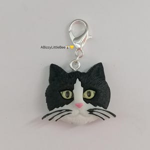May include: A black and white cat charm with green eyes and a pink nose. The charm is attached to a silver key ring.