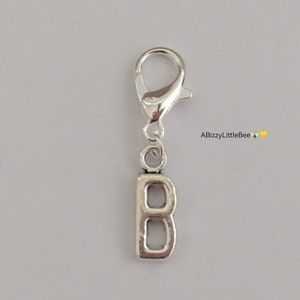 May include: Silver-coloured charm featuring a lobster clasp and a lowercase letter "b" pendant. The charm is likely made of metal and is designed to be attached to a bracelet, necklace, or other jewellery item. The background is a light grey colour.