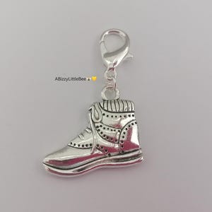 Peut inclure: Charm argenté en forme de basket avec un fermoir. Le charm présente des coutures et des perforations détaillées, et le texte "ABizzyLittleBee" est visible dans le coin supérieur gauche.