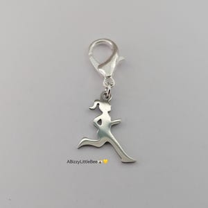 May include: A silver-toned metal charm featuring a silhouette of a person running, attached to a lobster claw clasp. The charm has a smooth, reflective surface. The text ABizzyLittleBee with a bee and heart emoji is printed below the charm.