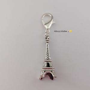 May include: A silver Eiffel Tower charm with a lobster clasp. The charm features a detailed design, with a black centre and red accents. The text "ABizzyLittleBee" is visible in the upper right corner.