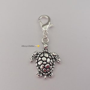 May include: A silver turtle charm with a lobster clasp. The charm has a detailed turtle design with a textured shell and flippers. The image includes the text "ABizzyLittleBee".