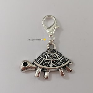 May include: Silver charm featuring a detailed turtle design. The turtle has a black shell and a silver body. The charm has a lobster claw clasp.