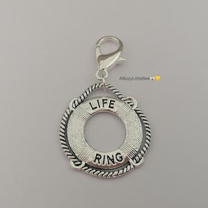May include: Silver-coloured charm in the shape of a life preserver ring, with the words "LIFE" and "RING" engraved on it. The charm is attached to a silver clasp. The charm is a nautical-themed accessory.