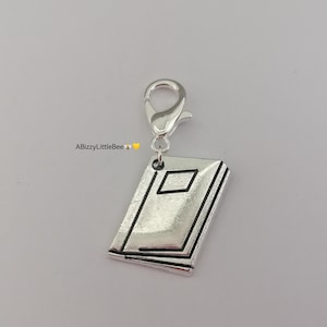 May include: A silver book charm with a rectangular design on the cover, attached to a lobster clasp. The text "ABizzyLittleBee" is in the upper left corner. The charm is made of metal.