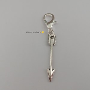 May include: A silver arrow charm with a lobster clasp. The arrow features a detailed fletching design and a pointed arrowhead. The charm is attached to a silver clasp, suitable for jewellery or accessories.