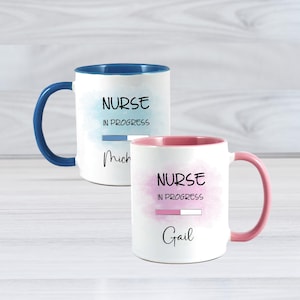 May include: Two white ceramic mugs with the text "NURSE IN PROGRESS". One mug has a blue interior and handle, the other has a pink interior and handle. The mugs have a watercolor design and the names "Mich" and "Gail" are written below the text.