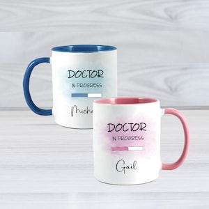 May include: Two white ceramic mugs with coloured handles and interiors. One has a blue handle and interior, the other a pink handle and interior. Both mugs feature the text "DOCTOR IN PROGRESS" with a progress bar and a name.