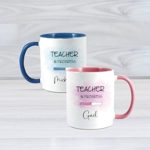 May include: Two white ceramic mugs with the text "TEACHER IN PROGRESS". One mug has a blue handle and interior, the other has a pink handle and interior. Each mug has a name below the text.