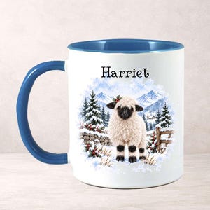 May include: A white ceramic mug with a blue handle and interior rim. The front features a graphic of a fluffy sheep with a black face standing in a snowy landscape with pine trees and mountains. The name Harriet is printed above the sheep.