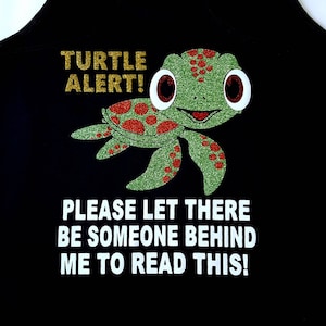 May include: Black tank top with a glittery green turtle graphic and the text "TURTLE ALERT!". Below the turtle is the text "PLEASE LET THERE BE SOMEONE BEHIND ME TO READ THIS!" in white.