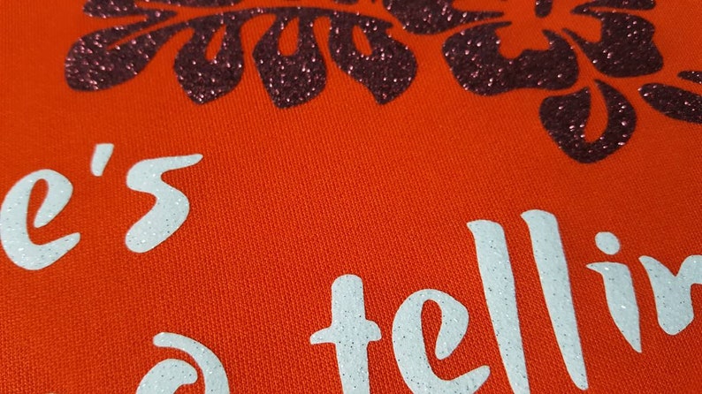 May include: Orange fabric with white glitter text that reads "She's telling" and a black glitter floral design.