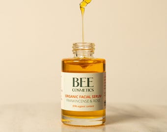 May include: A glass bottle of Bee Cosmetics Organic Facial Serum Frankincense & Rose. The bottle is filled with a golden yellow liquid and has a dropper attached. The bottle is sitting on a white surface.
