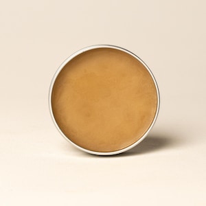 May include: A circular container with a silver rim, filled with a smooth, tan-coloured substance. The product is set against a neutral background, emphasising the texture and colour of the contents. The container's design is simple and clean.