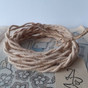 Puede incluir: Una bobina de hilo de yute natural, de color beige, descansa sobre papel vintage con motivos florales. El hilo está enrollado apretado, con un extremo suelto que se extiende desde la bobina. El fondo es azul claro.