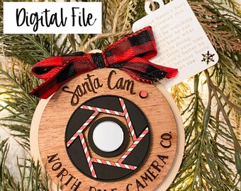 May include: A wooden Christmas ornament shaped like a camera lens with the words "Santa Cam North Pole Camera Co."  The ornament is decorated with red and white candy cane stripes and a red and black buffalo plaid bow. A white tag with text is attached to the ornament. The text on the tag reads: "Because Santa's busy, delivered this cam. I'll be watching whenever he can. You'll know what you're doing right from your tree. You'd better be good because Santa can see!"