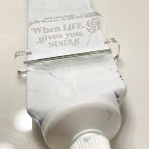 May include: A clear acrylic tube squeezer with the text "When LIFE gives you SUGAR" and a lollipop graphic. The squeezer is attached to a white tube of toothpaste.