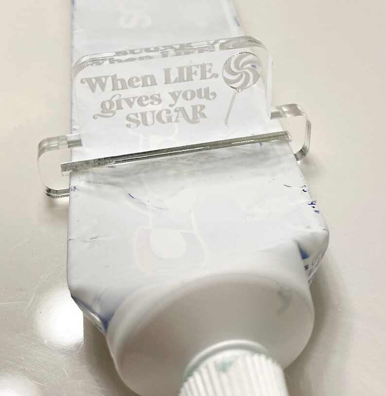 May include: A clear acrylic tube squeezer with the text "When LIFE gives you SUGAR" and a lollipop graphic. The squeezer is attached to a white tube of toothpaste.