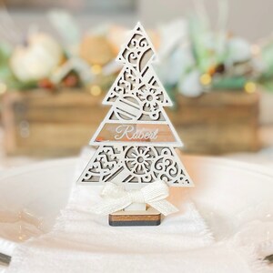 Puede incluir: Porta tarjetas de lugar de &aacute;rbol de Navidad de madera blanca con un lazo blanco y el nombre "Robert" escrito en &eacute;l.