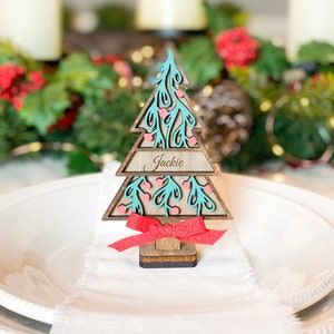 Puede incluir: Un marcador de lugar de &aacute;rbol de Navidad de madera con un dise&ntilde;o turquesa y rosa. El nombre "Jackie" est&aacute; escrito en la tarjeta.