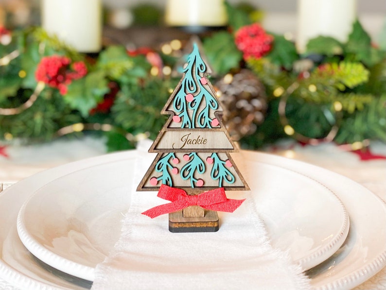 Puede incluir: Un marcador de lugar de &aacute;rbol de Navidad de madera con un dise&ntilde;o turquesa y rosa. El nombre "Jackie" est&aacute; escrito en la tarjeta.
