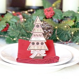 May include: A wooden Christmas tree place card with the word "Mom" on it. The tree is decorated with snowflakes and hearts. The place card is sitting on a red napkin on a white plate.