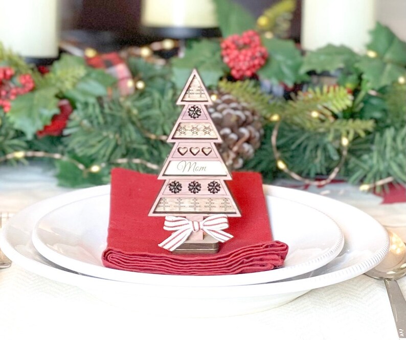 May include: A wooden Christmas tree place card with the word "Mom" on it. The tree is decorated with snowflakes and hearts. The place card is sitting on a red napkin on a white plate.