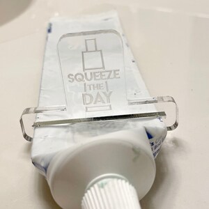 May include: A clear plastic toothpaste squeezer with the text "Squeeze the Day" printed on it. The squeezer is attached to a tube of toothpaste.