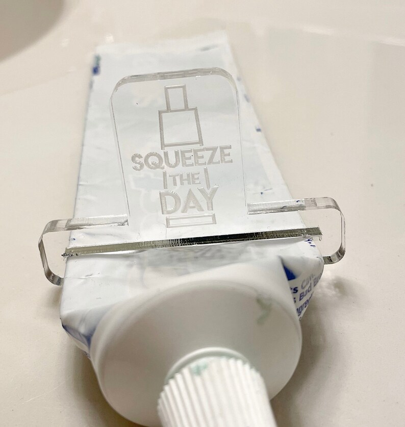 May include: A clear plastic toothpaste squeezer with the text "Squeeze the Day" printed on it. The squeezer is attached to a tube of toothpaste.