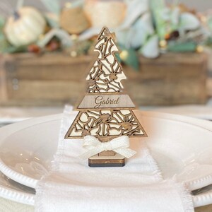 Puede incluir: Un marcador de lugar de &aacute;rbol de Navidad de madera con un dise&ntilde;o recortado y el nombre "Gabriel" grabado en &eacute;l. El &aacute;rbol est&aacute; decorado con un lazo de sat&eacute;n blanco y est&aacute; sentado en un plato blanco.