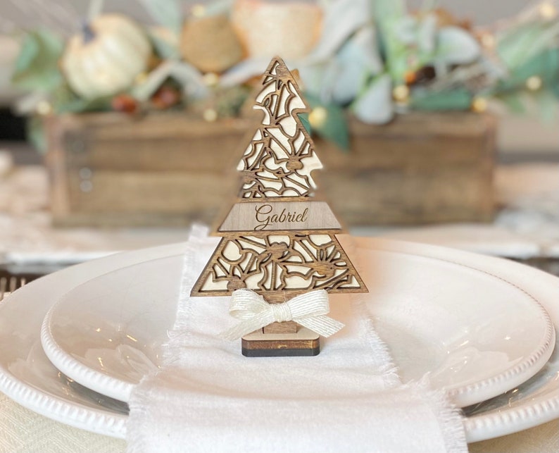 Puede incluir: Un marcador de lugar de &aacute;rbol de Navidad de madera con un dise&ntilde;o recortado y el nombre "Gabriel" grabado en &eacute;l. El &aacute;rbol est&aacute; decorado con un lazo de sat&eacute;n blanco y est&aacute; sentado en un plato blanco.