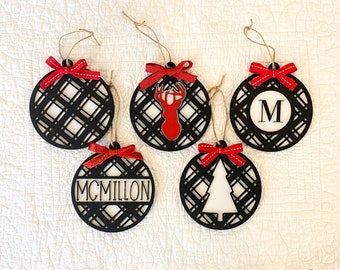 May include: Five black and white Christmas ornaments with red bows. The ornaments feature a plaid pattern, a deer, a tree, the letter M, and the name McMillon.