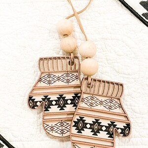 May include: Two wooden ornaments shaped like mittens with a black and white geometric pattern. The ornaments are attached to a brown cord with wooden beads.