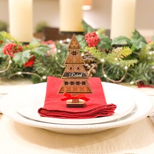 May include: A wooden Christmas tree place card with a red ribbon and the name "Robert" engraved on it. The place card is sitting on a red napkin on a white plate.