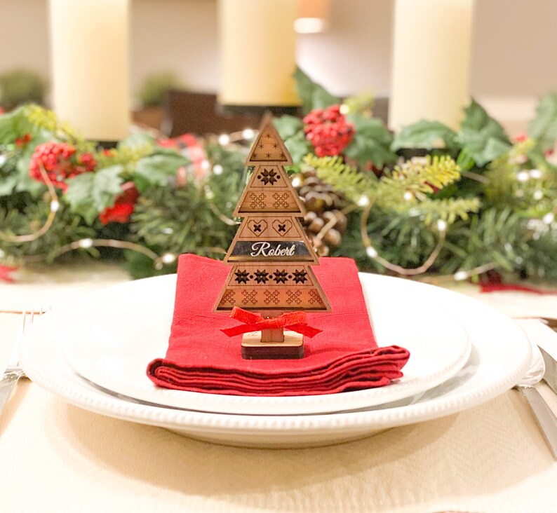May include: A wooden Christmas tree place card with a red ribbon and the name "Robert" engraved on it. The place card is sitting on a red napkin on a white plate.