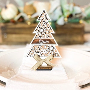 Puede incluir: Porta tarjetas de madera blanca en forma de &aacute;rbol de Navidad con un lazo dorado y el nombre "Gavin" en el nivel intermedio. El porta tarjetas est&aacute; sobre un plato blanco.