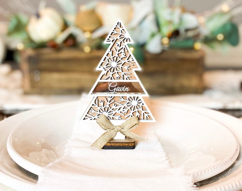 Puede incluir: Porta tarjetas de madera blanca en forma de &aacute;rbol de Navidad con un lazo dorado y el nombre "Gavin" en el nivel intermedio. El porta tarjetas est&aacute; sobre un plato blanco.
