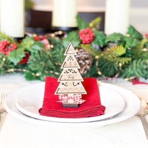 May include: A wooden Christmas tree place card with the name "Joey" engraved on it. The tree is decorated with a red and white gingham bow and is sitting on a red napkin on a white plate.