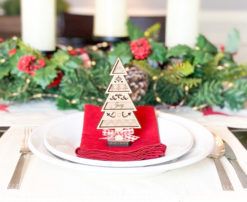 May include: A wooden Christmas tree place card with the name "Joey" engraved on it. The tree is decorated with a red and white gingham bow and is sitting on a red napkin on a white plate.