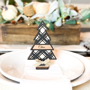 Puede incluir: Porta tarjetas de lugar de &aacute;rbol de Navidad de madera negra con una etiqueta de nombre marr&oacute;n que dice "Chloe". El porta tarjetas est&aacute; en un plato blanco con una servilleta blanca.