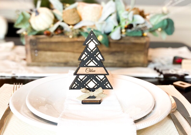 Puede incluir: Porta tarjetas de lugar de &aacute;rbol de Navidad de madera negra con una etiqueta de nombre marr&oacute;n que dice "Chloe". El porta tarjetas est&aacute; en un plato blanco con una servilleta blanca.