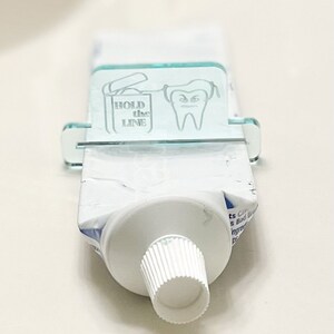 May include: A clear acrylic toothpaste dispenser with a tooth graphic and the text "HOLD the LINE". The dispenser is holding a tube of toothpaste.
