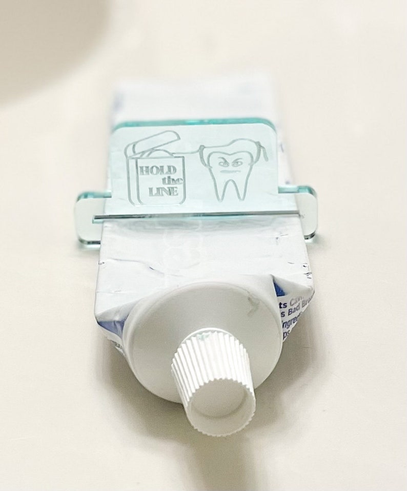 May include: A clear acrylic toothpaste dispenser with a tooth graphic and the text "HOLD the LINE". The dispenser is holding a tube of toothpaste.