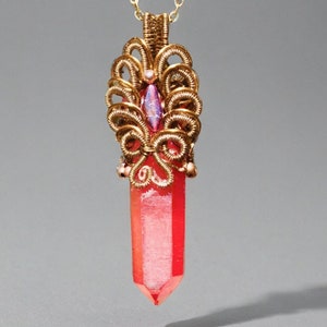 Può includere: Un ciondolo avvolto in filo d'oro con una punta di cristallo rosa. Il ciondolo presenta un design delicato e intricato con una piccola pietra preziosa iridescente nella parte superiore.