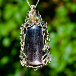 Puede incluir: Un colgante de alambre plateado envuelto alrededor de una piedra gris oscura con un patrón de remolino. El colgante está acentuado con pequeñas cuentas de plata y una piedra transparente en la parte superior.