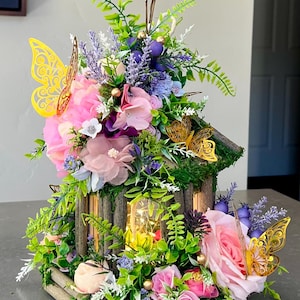 May include: A decorative wooden birdhouse adorned with pink and purple flowers, green foliage, and gold butterfly accents. The birdhouse has a moss-covered roof and contains a warm, glowing light inside. The overall design is whimsical and floral.
