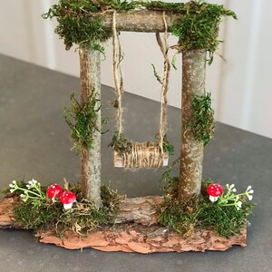 K&ouml;nnte beinhalten: Ein Miniatur-Schaukelgestell aus Holz mit Moos und kleinen roten Pilzen. Die Schaukel besteht aus Schnur und h&auml;ngt an zwei Holzpfosten. Das Schaukelgestell steht auf einem St&uuml;ck Rinde.